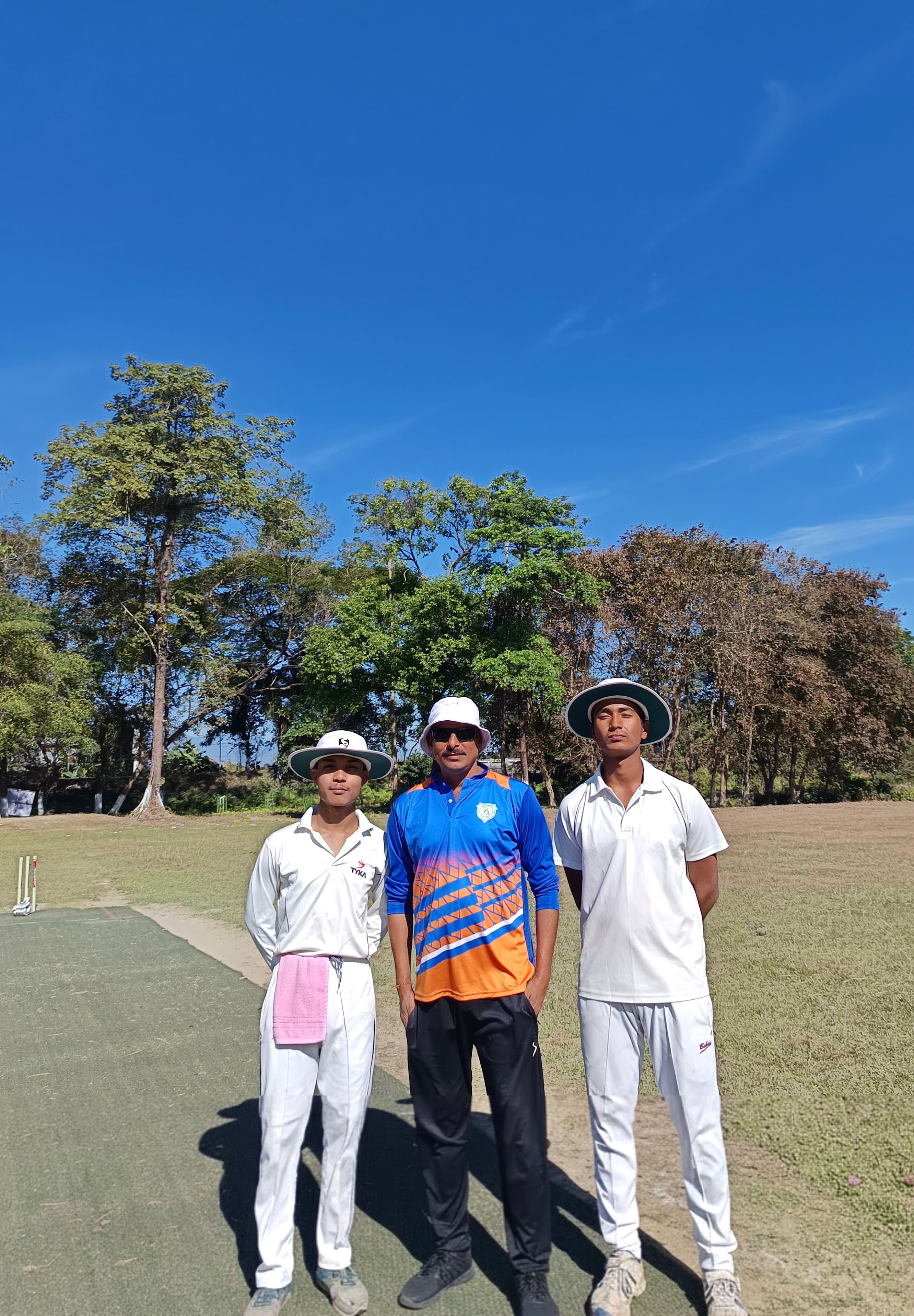 क्रिकेट की पोशाक पहने तीन पुरुष साफ नीले आसमान के नीचे मैदान पर खड़े हैं।