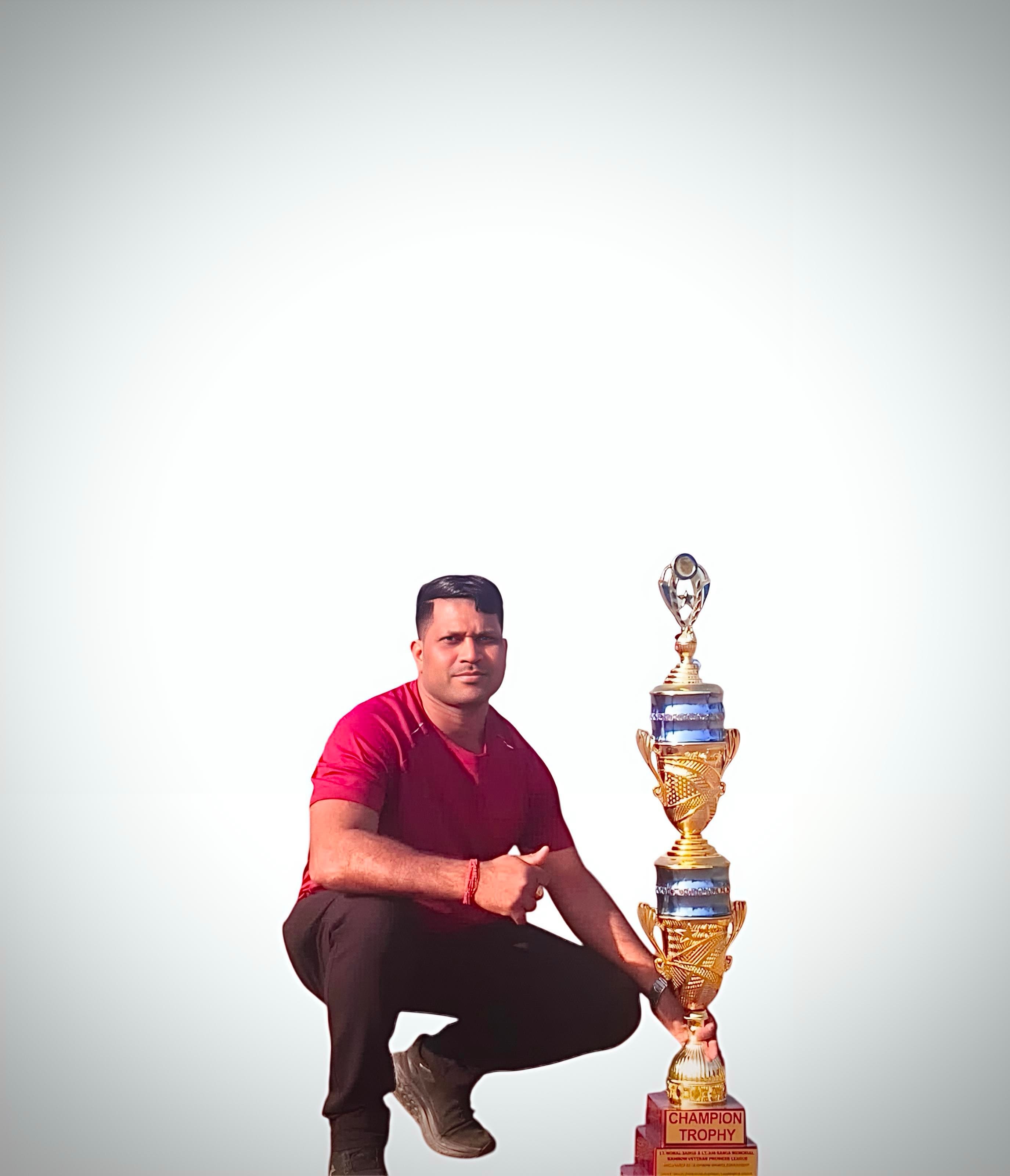 सफेद पृष्ठभूमि पर लंबी सुनहरी चैंपियन ट्रॉफी के पास बैठा लाल टी-शर्ट पहने व्यक्ति।