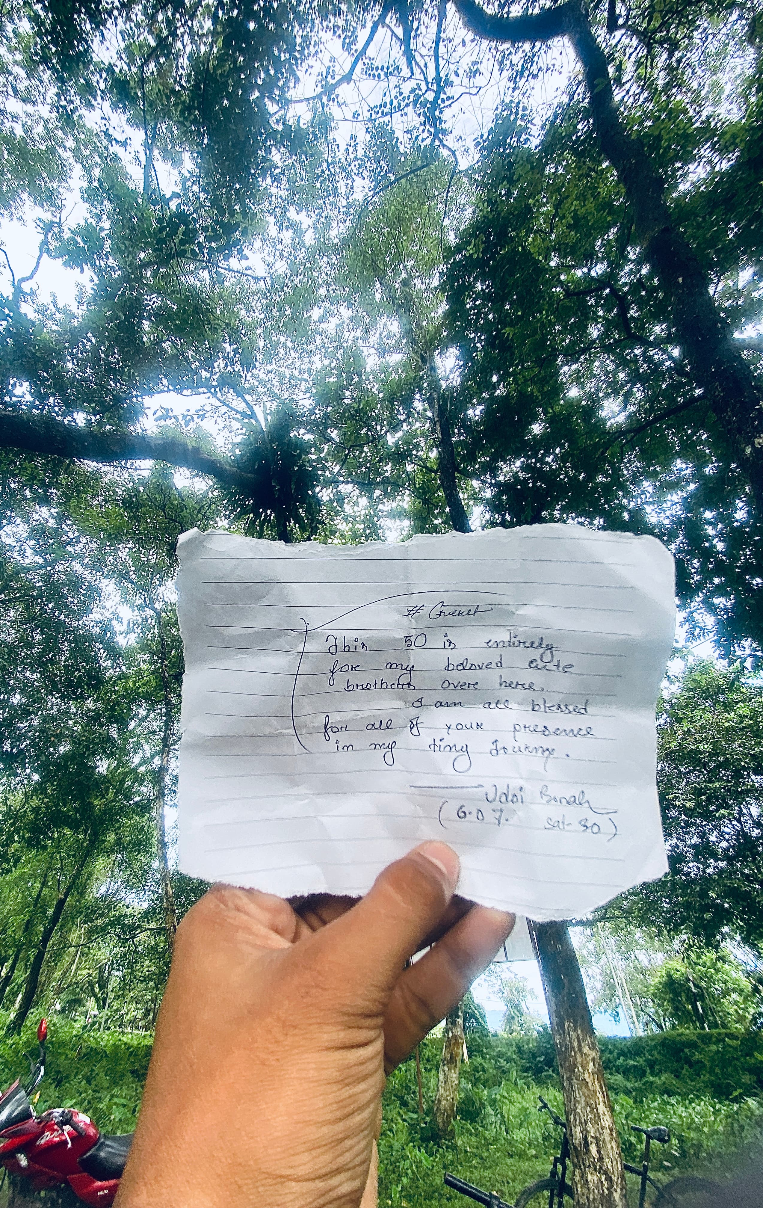 हरे-भरे पेड़ों की पृष्ठभूमि में एक हाथ में हस्तलिखित संदेश वाला कागज का टुकड़ा।