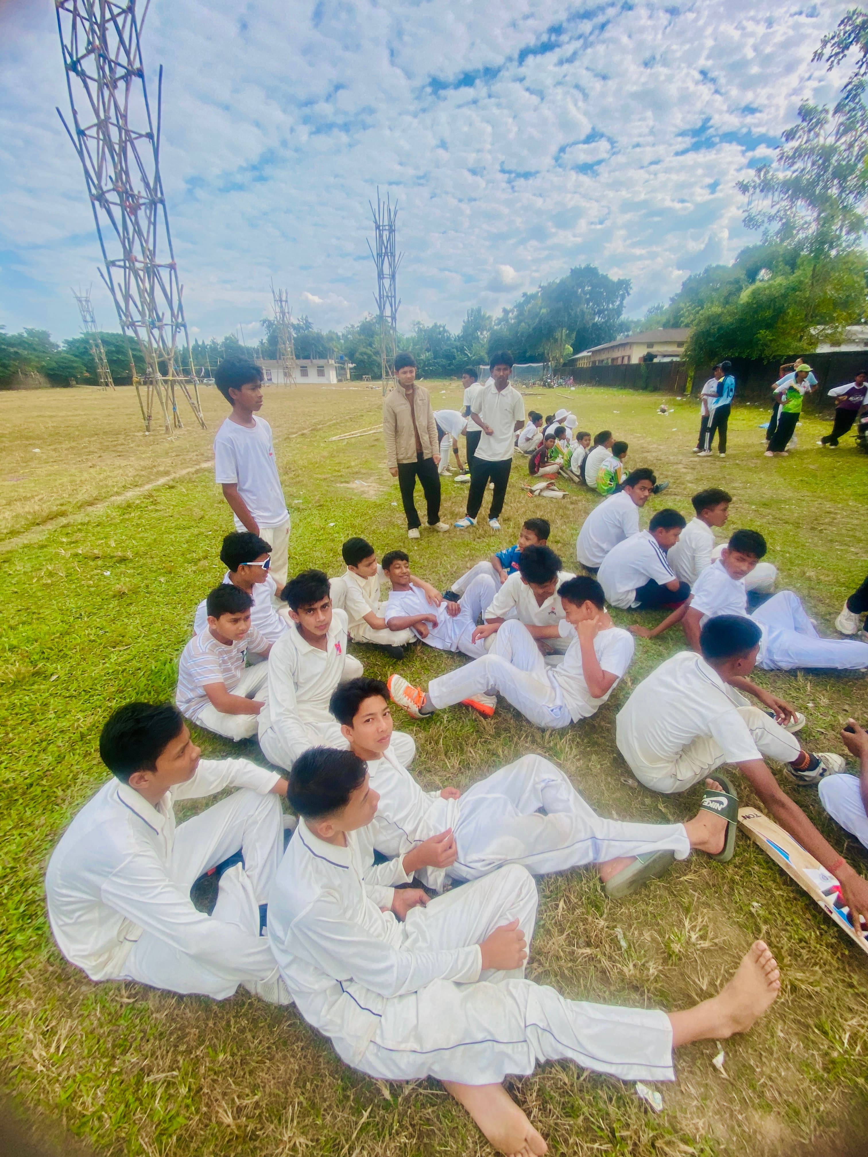 सफेद क्रिकेट वर्दी पहने लड़कों का समूह घास के मैदान पर बैठा है।