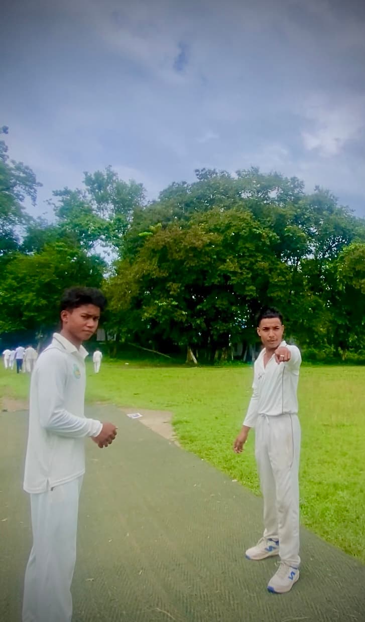 सफेद क्रिकेट वर्दी में दो युवक, एक इशारा करते हुए, पीछे मैदान और घने पेड़।